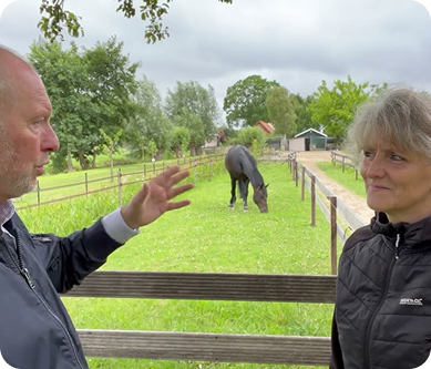 Iris and a man standing outdoors in a grassy area with a horse in the background Iris and a man standing outdoors in a grassy area with a horse in the background